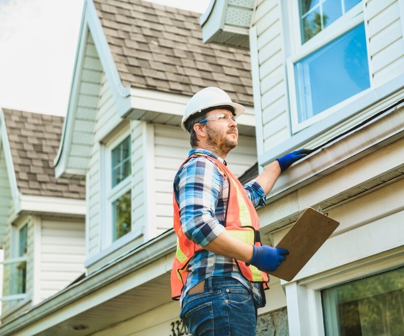 A construction worker in a hard hat and safety vest inspects the exterior of a house, holding a clipboard - Roofing Materials
