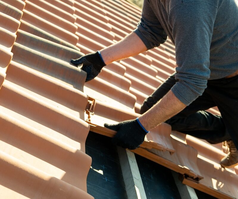 A person wearing gloves adjusts a red roof tile while working on a roof repair - Roof shingles