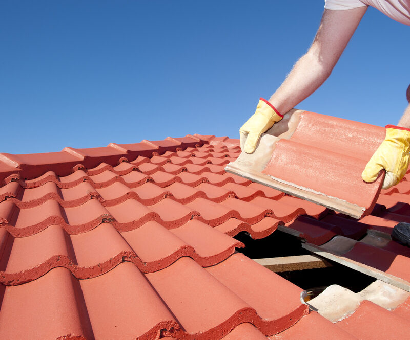 A worker wearing gloves replaces a damaged roof tile on a red-tiled roof under a clear blue sky - tile roof