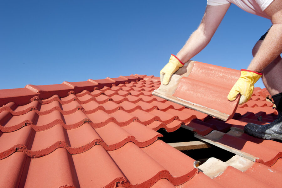 A worker wearing gloves replaces a damaged roof tile on a red-tiled roof under a clear blue sky - tile roof